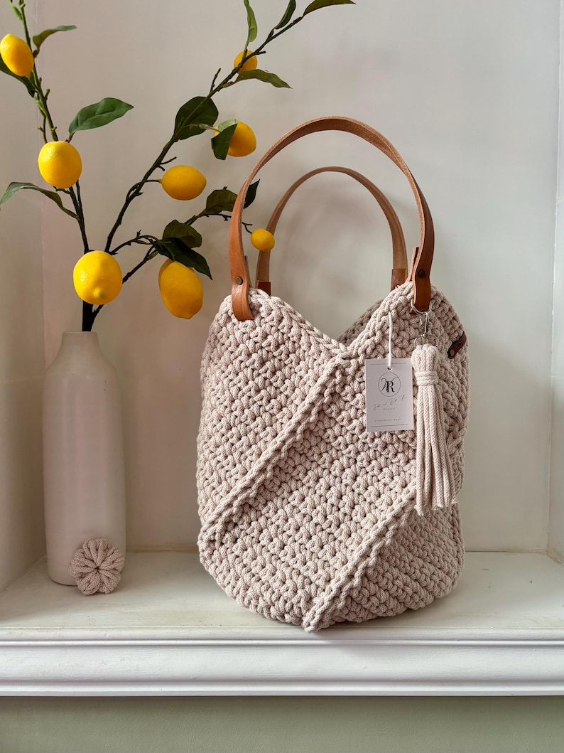 May include: A beige crochet handbag featuring a textured knit pattern, two tan leather handles, and a matching tassel charm. A small white rectangular tag with the letter R is attached to the handle. The bag has a structured, rounded shape.