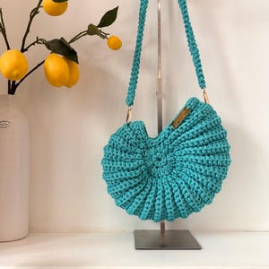 May include: A round, turquoise crochet handbag with a textured, ribbed spiral pattern. The bag features a matching braided shoulder strap attached with gold-toned metal hardware and a small rectangular gold-toned metal accent on the top edge.