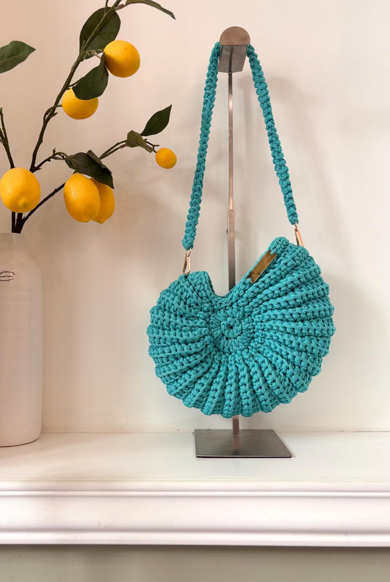 May include: A round, turquoise crochet handbag with a textured, ribbed spiral pattern. The bag features a matching braided shoulder strap attached with gold-toned metal hardware and a small rectangular gold-toned metal accent on the top edge.