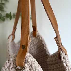 May include: A beige crocheted tote bag featuring thick, light brown leather straps. The straps are securely attached to the textured, woven body of the bag with metallic rivets and silver-toned metal rings, creating a sturdy and functional design.