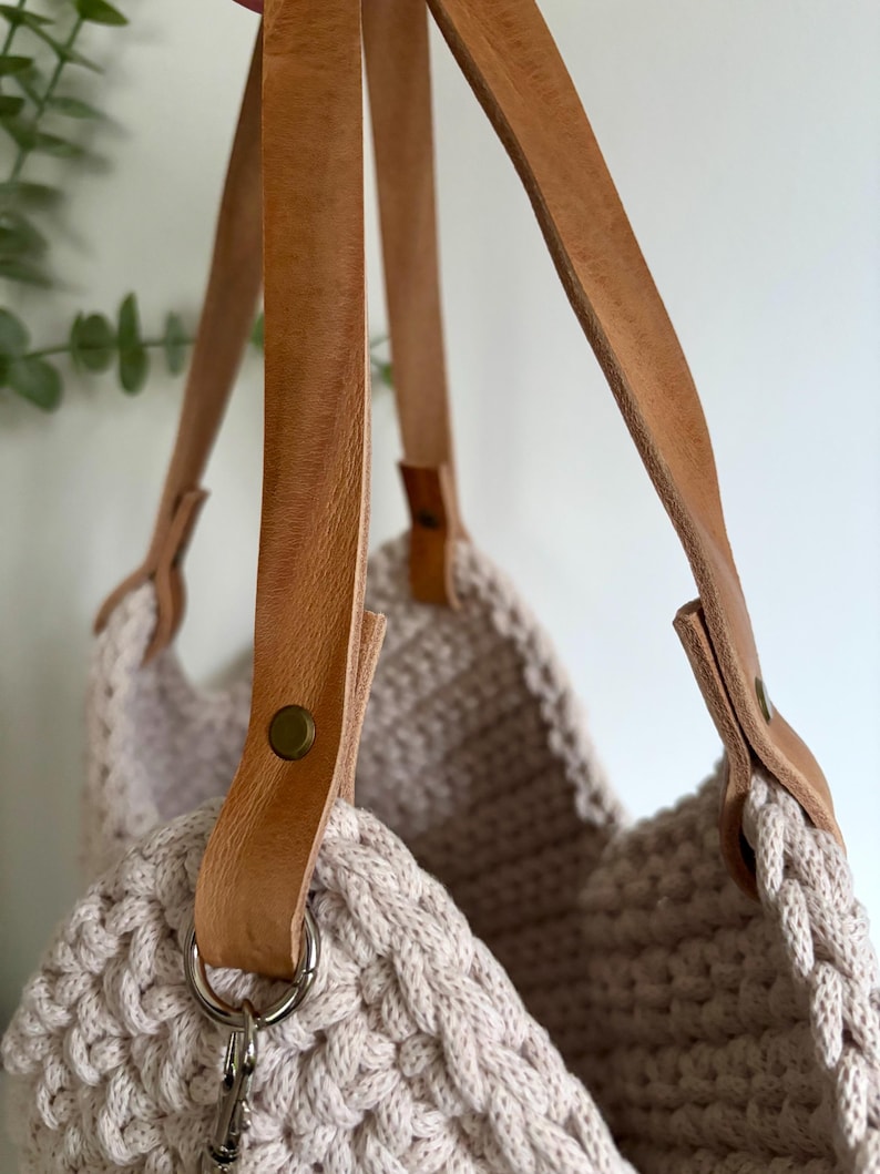 May include: A beige crocheted tote bag featuring thick, light brown leather straps. The straps are securely attached to the textured, woven body of the bag with metallic rivets and silver-toned metal rings, creating a sturdy and functional design.