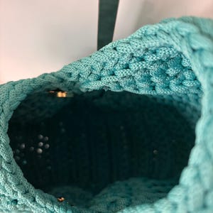 May include: A turquoise crochet bag with a thick, textured cord construction. The interior is visible, showing a deep, dark teal lining. A small, metallic gold-toned hardware piece is attached to the exterior rim, providing a subtle contrast.