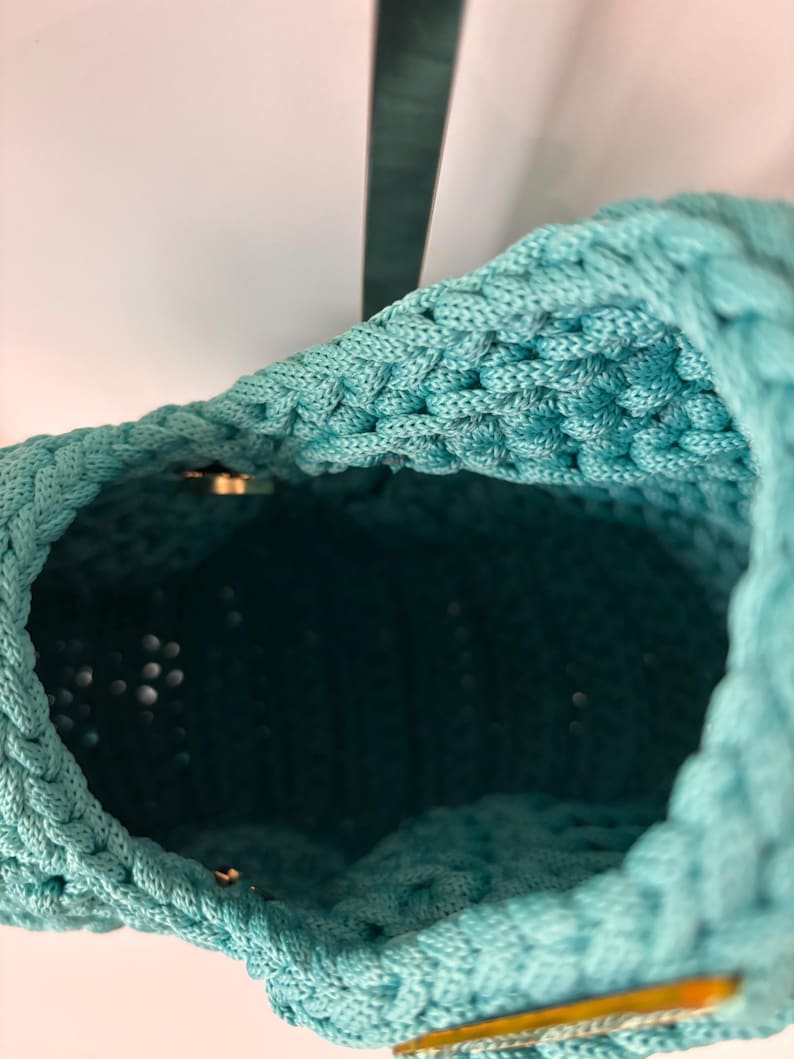 May include: A turquoise crochet bag with a thick, textured cord construction. The interior is visible, showing a deep, dark teal lining. A small, metallic gold-toned hardware piece is attached to the exterior rim, providing a subtle contrast.