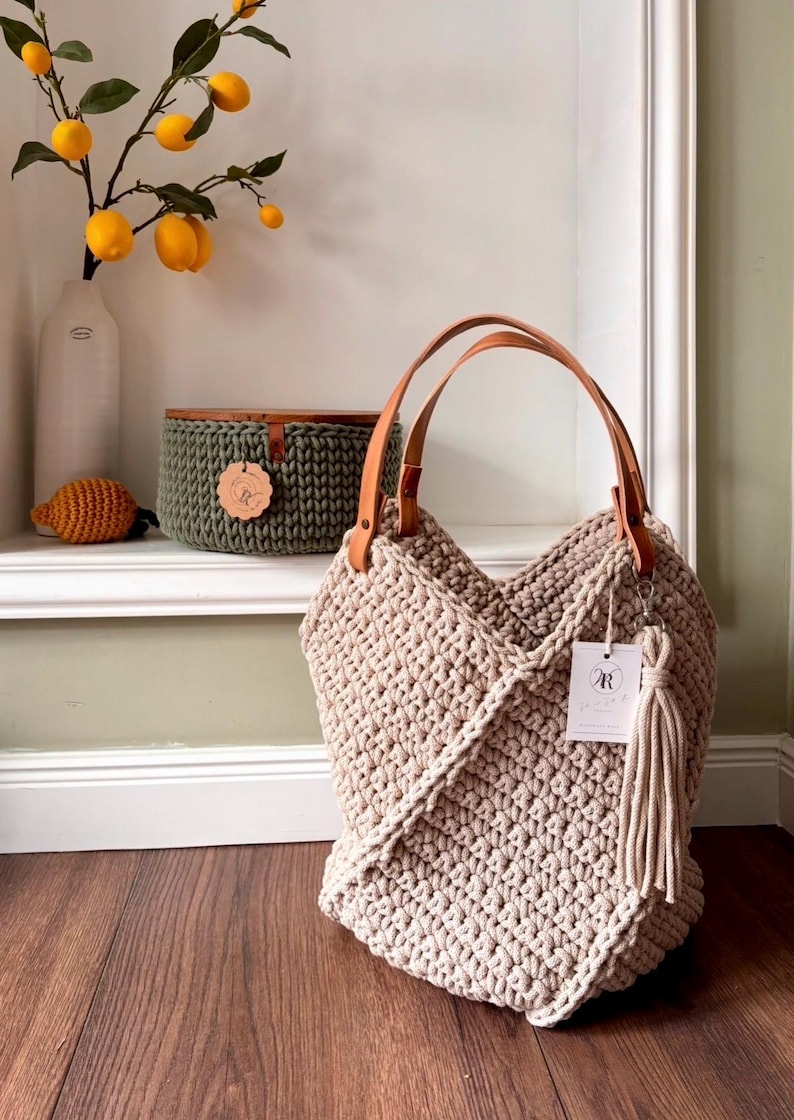 May include: A beige crochet tote bag featuring a textured knit pattern, two tan leather handles, and a matching beige tassel hanging from a silver clasp. A white rectangular tag with the letter R is attached to the side of the bag.