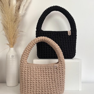 Könnte beinhalten: Zwei gehäkelte Handtaschen, eine schwarze und eine beige, mit einem einzigen Henkel und einem kleinen Lederanhänger. Die Taschen werden auf einer weißen Oberfläche präsentiert.