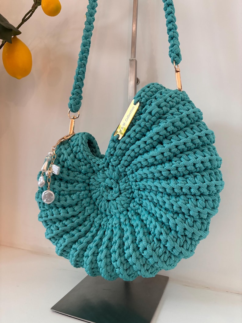 Op de afbeelding: Een blauwgroene gehaakte handtas met een spiraalvormig schelpontwerp, voorzien van een dikke gevlochten schouderband, goudkleurige hardware en een bedel met witte parels. Een klein gouden metalen label bevat de tekst W.&W. Collection.
