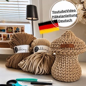 Könnte beinhalten: Zwei Knäuel beiges Bobbiny-Garn, ein gehäkeltes pilzförmiges Gefäß mit braunem Lederetikett, eine türkise Häkelnadel, eine kleine schwarze Häkelnadel und eine Schere mit türkisen Griffen, inklusive deutscher Flagge und Text.