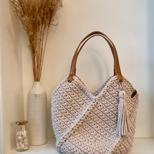 May include: A beige crochet tote bag featuring a textured stitch pattern, two tan faux leather handles attached with metal rivets, and a matching beige fabric tassel hanging from one of the handle rings on the side of the bag.