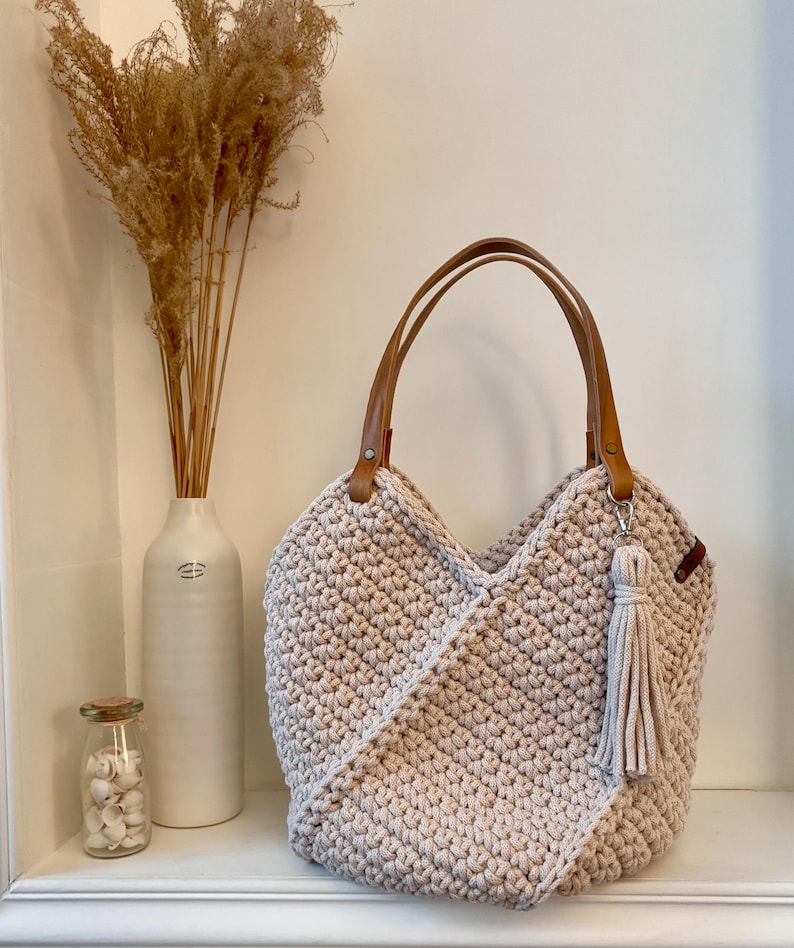 May include: A beige crochet tote bag featuring a textured stitch pattern, two tan faux leather handles attached with metal rivets, and a matching beige fabric tassel hanging from one of the handle rings on the side of the bag.