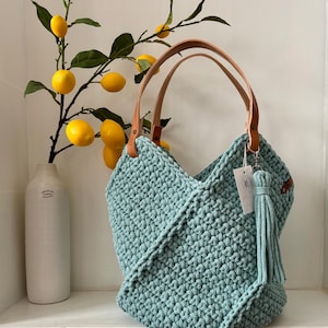 K&ouml;nnte beinhalten: Eine salbeigr&uuml;ne H&auml;keltasche mit strukturiertem Knotenmuster, zwei hellbraunen Lederhenkeln und einer passenden Garnquaste an einem silbernen Metallring. Ein kleines wei&szlig;es rechteckiges Etikett mit einem kreisf&ouml;rmigen Logo ist am Henkel befestigt.