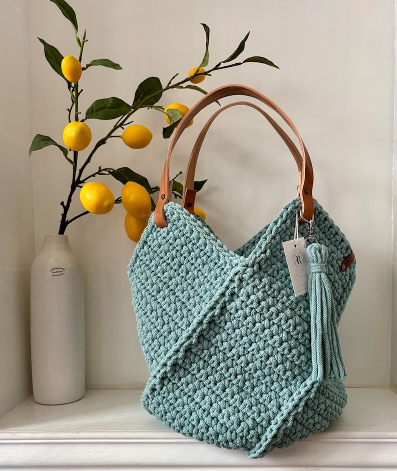 K&ouml;nnte beinhalten: Eine salbeigr&uuml;ne H&auml;keltasche mit strukturiertem Knotenmuster, zwei hellbraunen Lederhenkeln und einer passenden Garnquaste an einem silbernen Metallring. Ein kleines wei&szlig;es rechteckiges Etikett mit einem kreisf&ouml;rmigen Logo ist am Henkel befestigt.