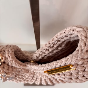 Op de afbeelding: Een beige gehaakte handtas met een dikke gevlochten rand en een goudkleurig metalen label met de tekst W & W Collection. De tas is voorzien van een ronde goudkleurige magnetische sluiting en een decoratieve hanger met doorzichtige kralen.