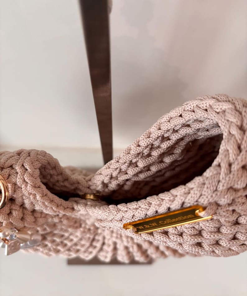 Op de afbeelding: Een beige gehaakte handtas met een dikke gevlochten rand en een goudkleurig metalen label met de tekst W & W Collection. De tas is voorzien van een ronde goudkleurige magnetische sluiting en een decoratieve hanger met doorzichtige kralen.