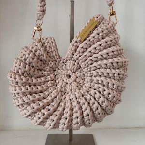 Op de afbeelding: Een beige gehaakte handtas met een cirkelvormig, schelpachtig ontwerp en een gevlochten schouderband. De tas is voorzien van goudkleurige metalen hardware, waaronder karabijnhaken en een rechthoekig label met de tekst W.&W. Collection.