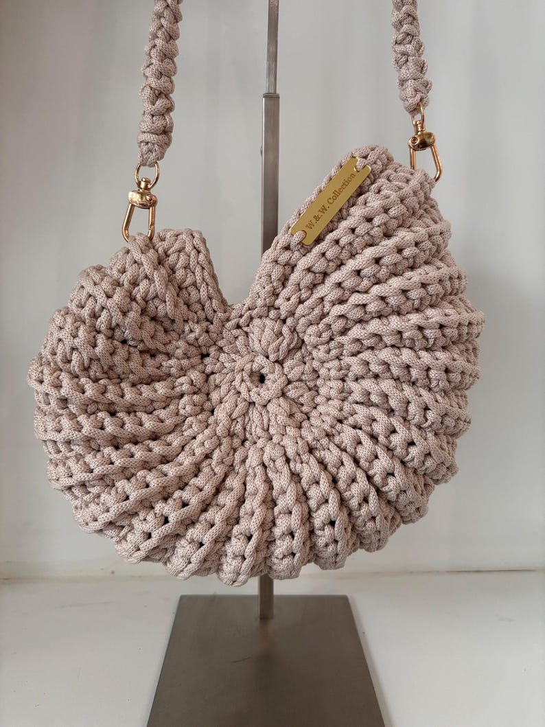 Op de afbeelding: Een beige gehaakte handtas met een cirkelvormig, schelpachtig ontwerp en een gevlochten schouderband. De tas is voorzien van goudkleurige metalen hardware, waaronder karabijnhaken en een rechthoekig label met de tekst W.&W. Collection.