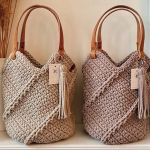K&ouml;nnte beinhalten: Zwei geh&auml;kelte Tragetaschen mit strukturiertem Diagonalmuster, eine in hellem Beige und eine in Mauve. Jede Tasche hat zwei hellbraune Ledergriffe, die mit Metallnieten befestigt sind, und eine passende Garnquaste an einem silberfarbenen Verschluss.