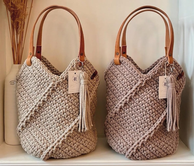 K&ouml;nnte beinhalten: Zwei geh&auml;kelte Tragetaschen mit strukturiertem Diagonalmuster, eine in hellem Beige und eine in Mauve. Jede Tasche hat zwei hellbraune Ledergriffe, die mit Metallnieten befestigt sind, und eine passende Garnquaste an einem silberfarbenen Verschluss.