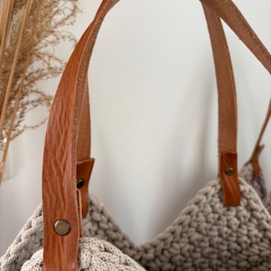 Könnte beinhalten: Beige gehäkelte Tragetasche mit braunen Ledergriffen. Die Textur der Tasche ist sichtbar, und die Lederriemen sind mit messingfarbenen Nieten befestigt. Die Tasche ist mit einer breiten Öffnung und einer stabilen Konstruktion versehen.