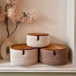 Könnte beinhalten: Drei runde Aufbewahrungskörbe aus Häkelarbeit mit Holzdeckeln. Die Körbe sind in Creme- und Brauntönen gehalten und haben einen Lederakzent auf dem Deckel. Die Körbe sind unterschiedlich groß und werden auf einem weißen Regal ausgestellt.