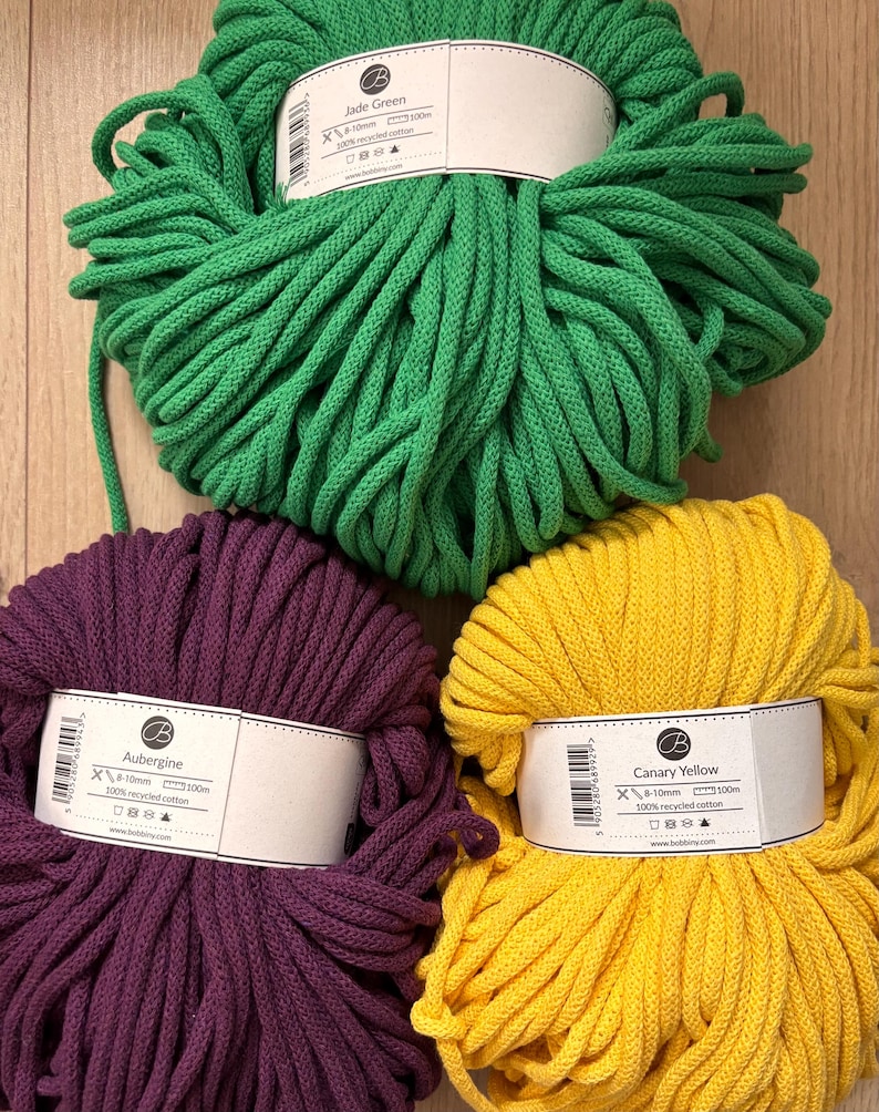 K&ouml;nnte beinhalten: Drei Garnkn&auml;uel aus geflochtener Kordel in Jadegr&uuml;n, Aubergine und Kanariengelb. Jedes Kn&auml;uel hat ein wei&szlig;es Papieretikett mit der Farbbezeichnung, 8-10mm St&auml;rke, 100m L&auml;nge und dem Hinweis auf 100% recycelte Baumwolle auf der Vorderseite.