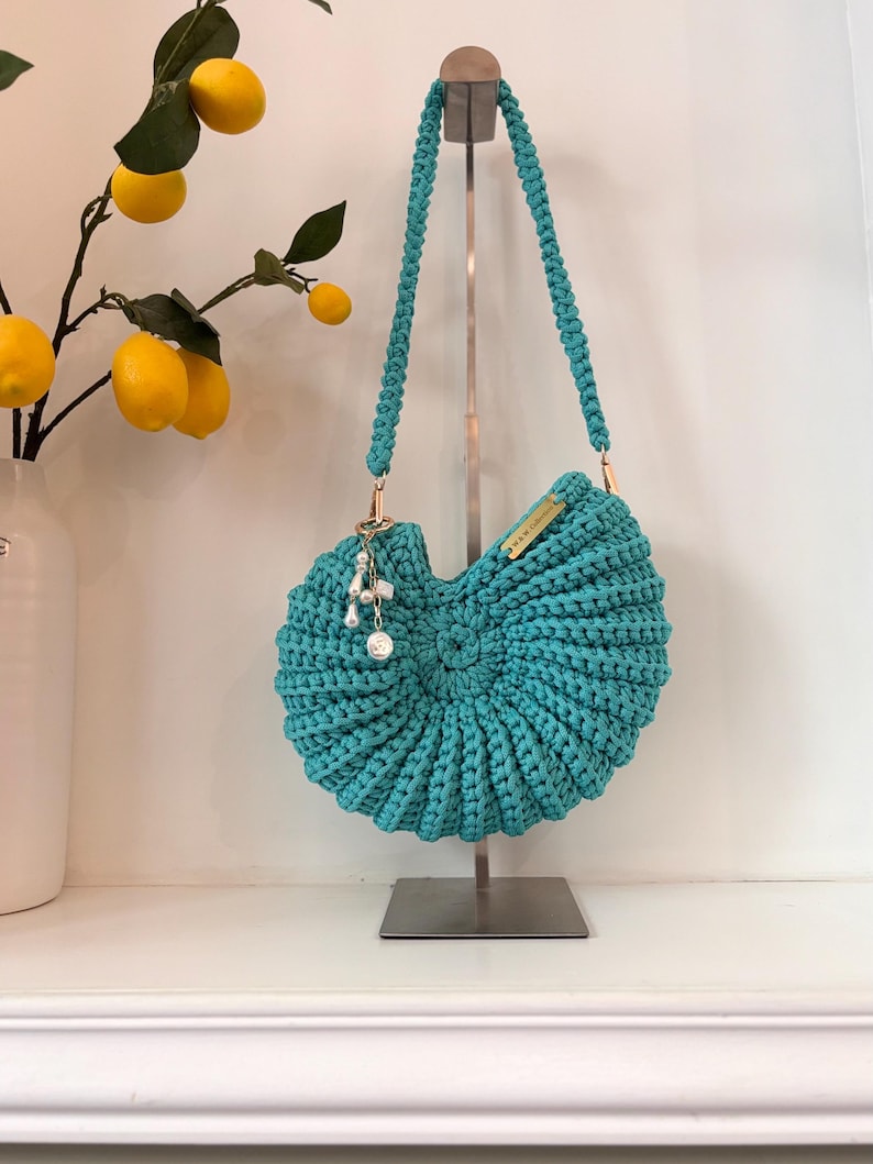 Op de afbeelding: Een tealgekleurde gehaakte handtas met een spiraalvormig schelpontwerp en een enkele gevlochten schouderband. De tas is voorzien van een goudkleurig metalen label met de tekst W&W Collection en een bedel met witte parels en een metalen hanger.