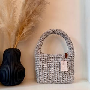 Könnte beinhalten: Eine hellgraue, handgehäkelte Tragetasche mit rundem Griff und rechteckigem Körper. Die Tasche hat ein kleines braunes Lederetikett und ein weißes Produktetikett. Die Tasche wird auf einem weißen Regal neben einer schwarzen Vase mit getrocknetem Pampasgras ausgestellt.
