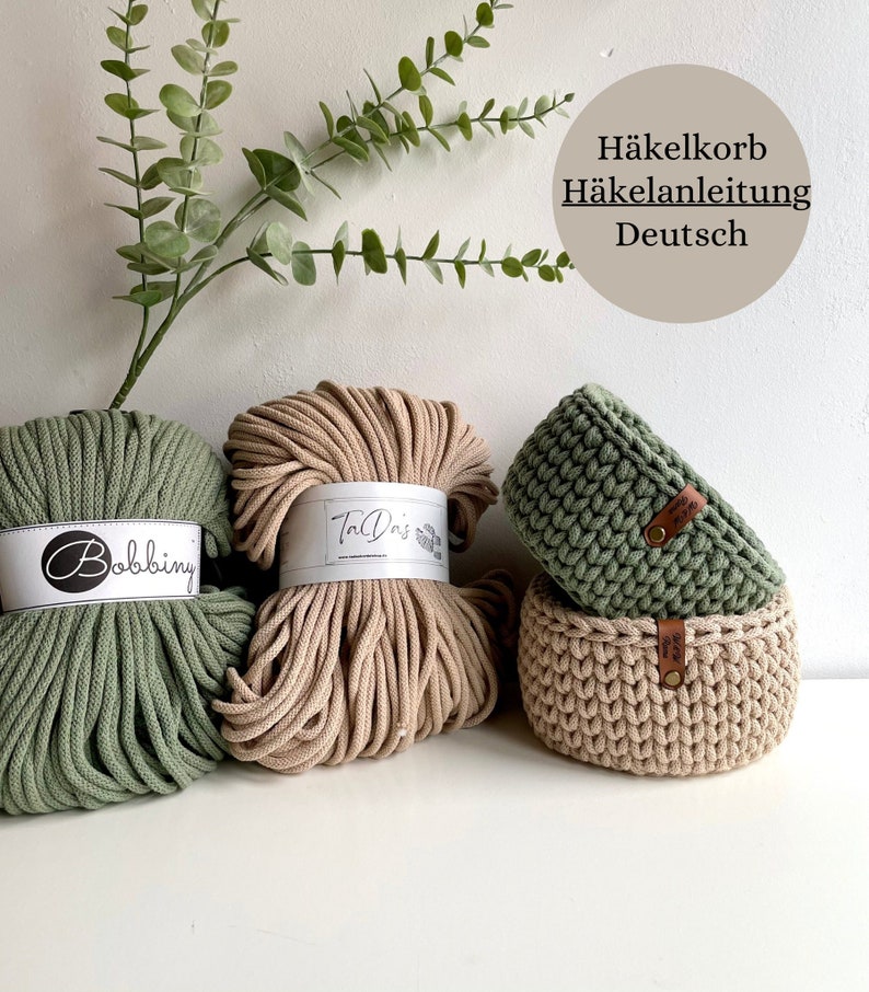 Puede incluir: Dos ovillos de cuerda de algod&oacute;n, uno verde y otro beige, con etiquetas que dicen "Bobbiny" y "The Oris". Dos cestas de ganchillo, una verde y otra beige, con etiquetas de cuero. El texto "H&auml;kelkorb H&auml;kelanleitung Deutsch" es visible en la imagen.