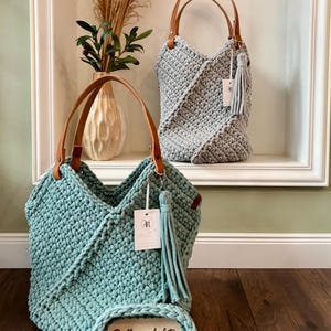 May include: Two handmade crochet bags with tan leather handles and matching tassels. One bag is light blue, the other is light gray. The light blue bag features a small oval tag with the text Brillenparkplatz and a white brand label.