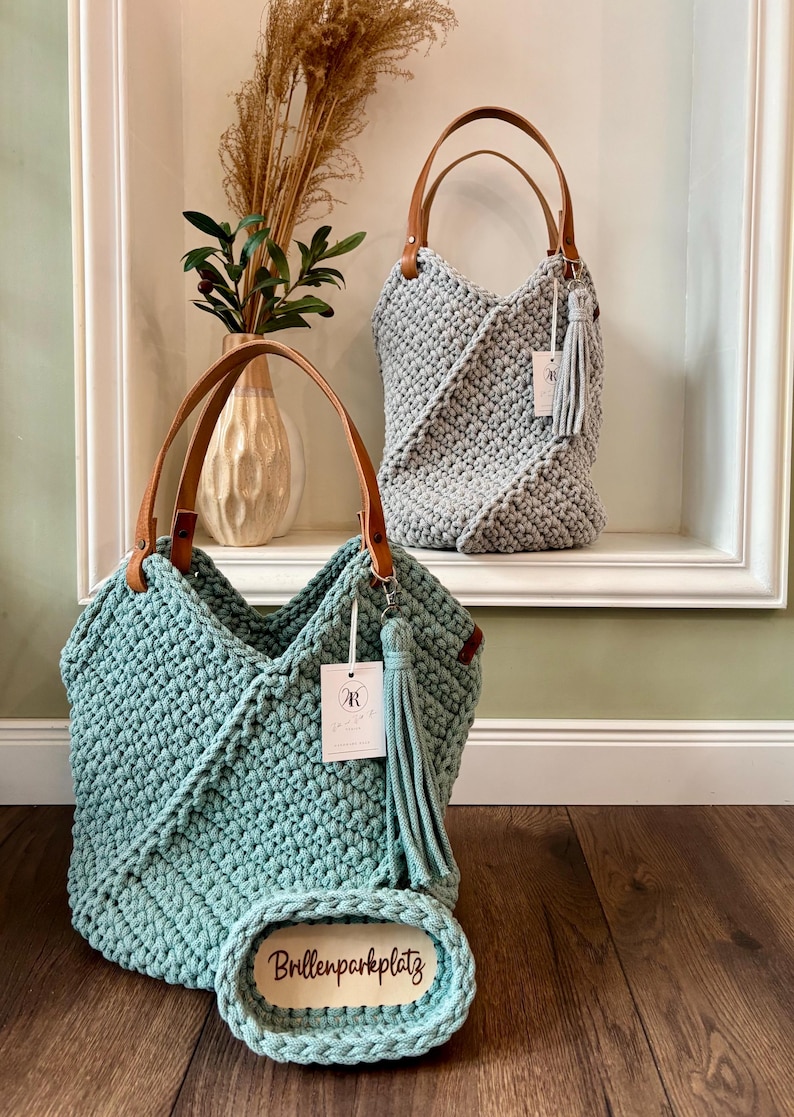 May include: Two handmade crochet bags with tan leather handles and matching tassels. One bag is light blue, the other is light gray. The light blue bag features a small oval tag with the text Brillenparkplatz and a white brand label.