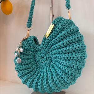 May include: A teal crochet handbag with a shell-like spiral design, featuring a thick braided shoulder strap, gold-tone hardware, and a dangling charm with white pearls. A small gold metal tag is attached, engraved with the text W.&W. Collection.