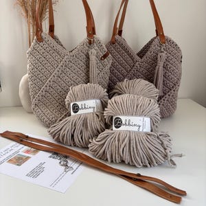 K&ouml;nnte beinhalten: Zwei geh&auml;kelte Handtaschen in Beige und Taupe mit braunen Ledergriffen und passenden Quasten, begleitet von zwei Kn&auml;ueln Bobbiny 5mm geflochtener Kordel, einem langen braunen Lederriemen mit Metallbeschl&auml;gen und einer Anleitungskarte aus Papier.