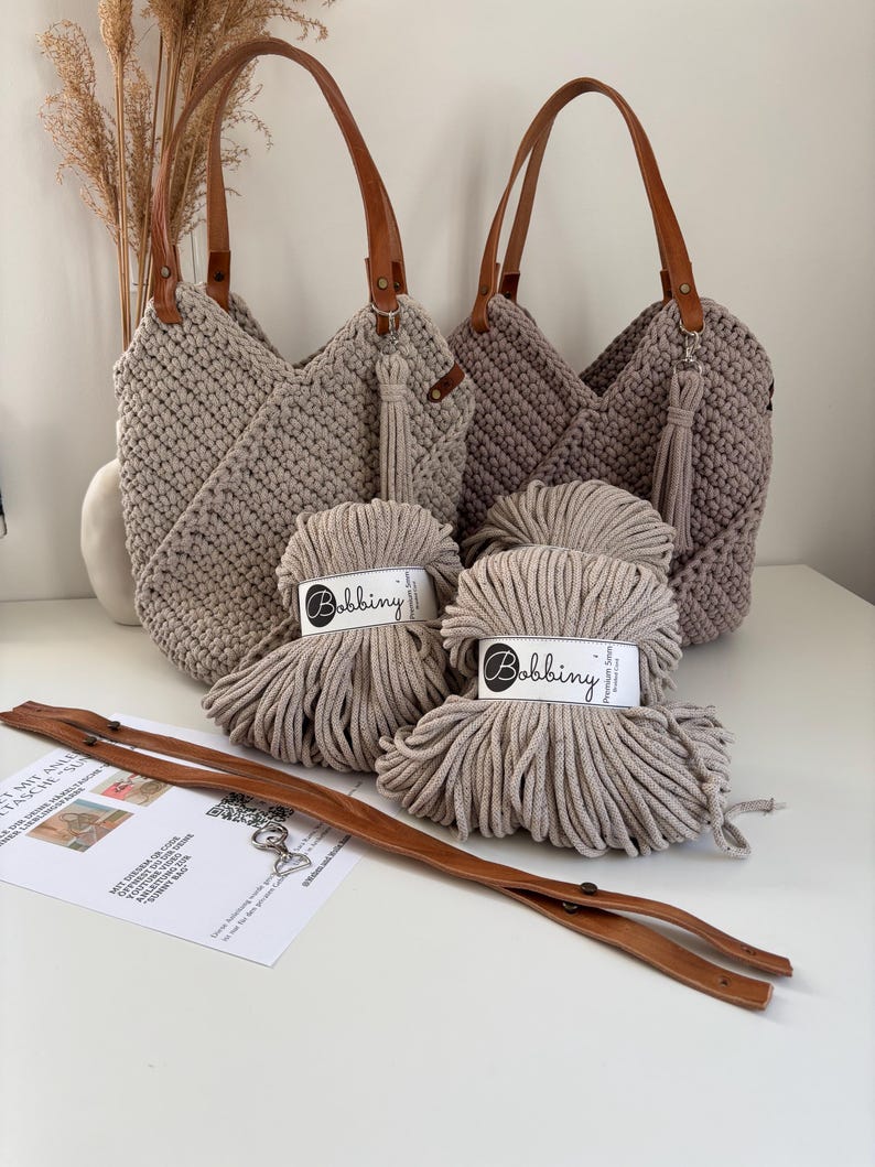K&ouml;nnte beinhalten: Zwei geh&auml;kelte Handtaschen in Beige und Taupe mit braunen Ledergriffen und passenden Quasten, begleitet von zwei Kn&auml;ueln Bobbiny 5mm geflochtener Kordel, einem langen braunen Lederriemen mit Metallbeschl&auml;gen und einer Anleitungskarte aus Papier.