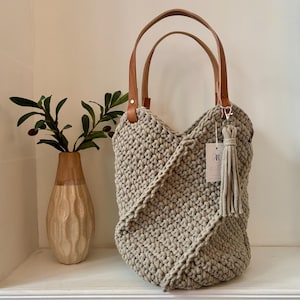 May include: A beige crocheted tote bag with two tan leather handles and a matching beige tassel attached to a silver clasp. A small white rectangular tag with the letter R is attached to the bag. The bag features a textured, knotted stitch pattern.