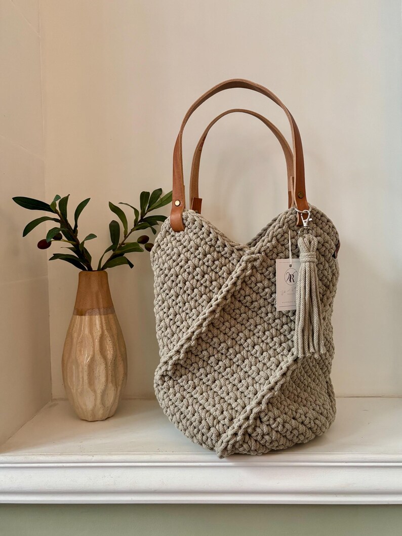 May include: A beige crocheted tote bag with two tan leather handles and a matching beige tassel attached to a silver clasp. A small white rectangular tag with the letter R is attached to the bag. The bag features a textured, knotted stitch pattern.