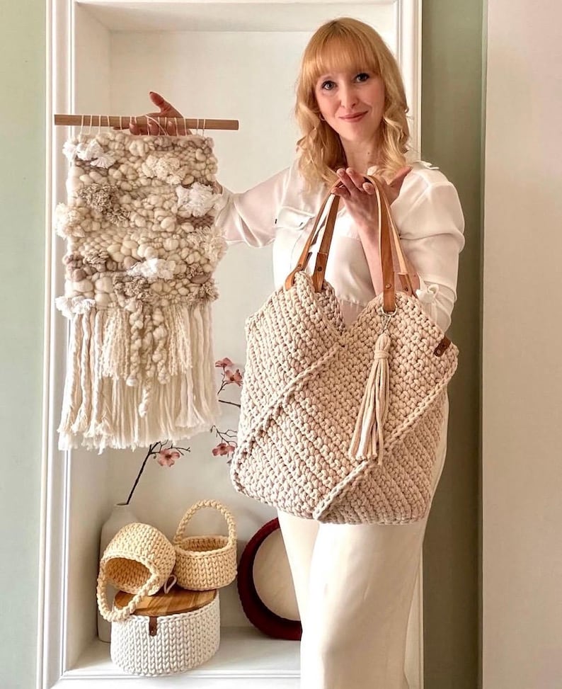 May include: A beige crocheted tote bag with leather handles and a matching tassel, a rectangular wall hanging with textured cream and tan yarn, two small round crocheted baskets, and a larger round crocheted basket with a wooden lid.