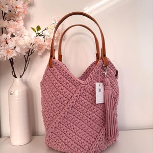 May include: A dusty rose crochet handbag featuring a textured knit pattern, two tan leather handles attached with metal rivets, and a matching long yarn tassel hanging from a silver lobster clasp. A small white rectangular tag is attached to the bag.