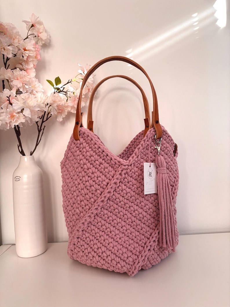 May include: A dusty rose crochet handbag featuring a textured knit pattern, two tan leather handles attached with metal rivets, and a matching long yarn tassel hanging from a silver lobster clasp. A small white rectangular tag is attached to the bag.