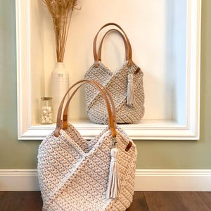 May include: Two beige crochet handbags featuring a textured stitch pattern, tan leather handles, and a matching beige tassel attached to a silver-toned metal clasp. The bags have a structured, angular design with a wide opening and a thick, durable weave.