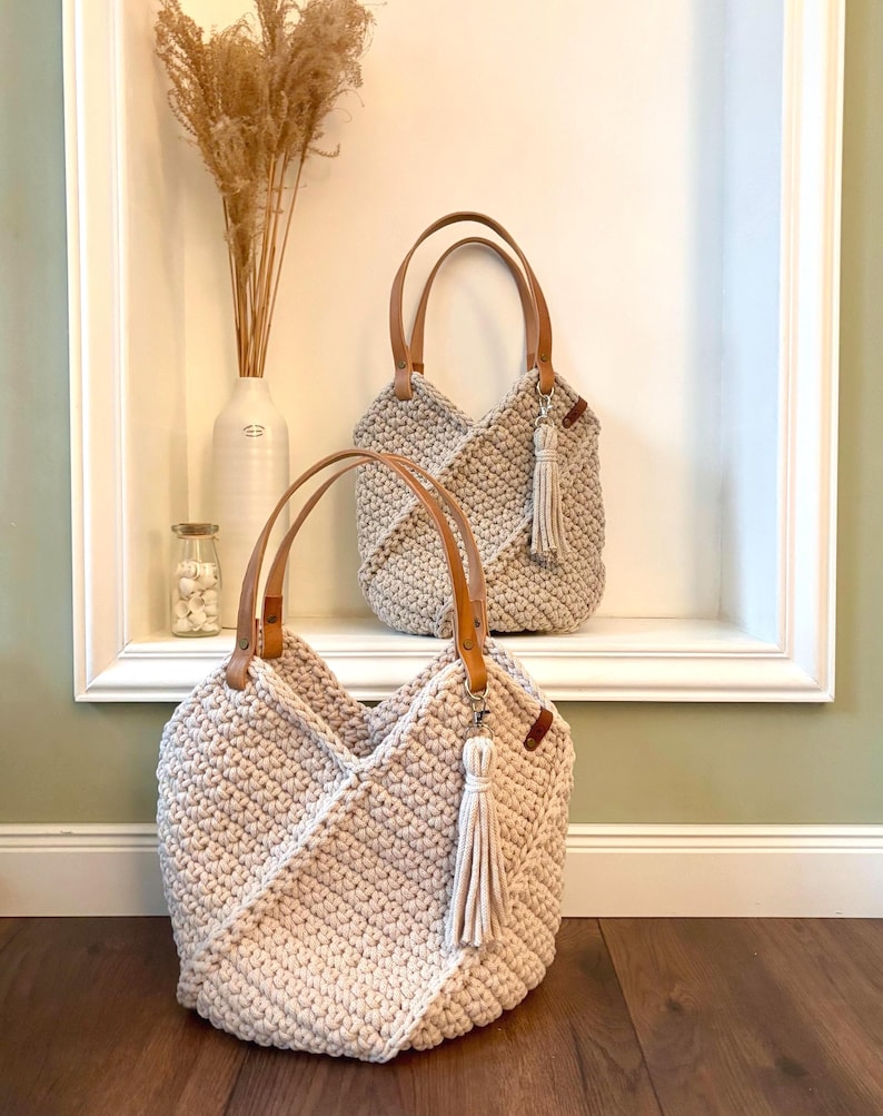 May include: Two beige crochet handbags featuring a textured stitch pattern, tan leather handles, and a matching beige tassel attached to a silver-toned metal clasp. The bags have a structured, angular design with a wide opening and a thick, durable weave.