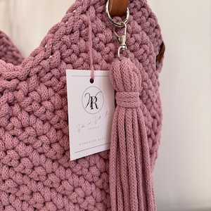 May include: A mauve crocheted handbag featuring a thick, textured knit pattern. The bag includes a brown leather strap attached with a silver metal clasp. A long, matching mauve cord tassel hangs from the clasp, and a white rectangular tag with the logo WR and the text "Willa and Willa Rose Design Handmade Bags" is attached.