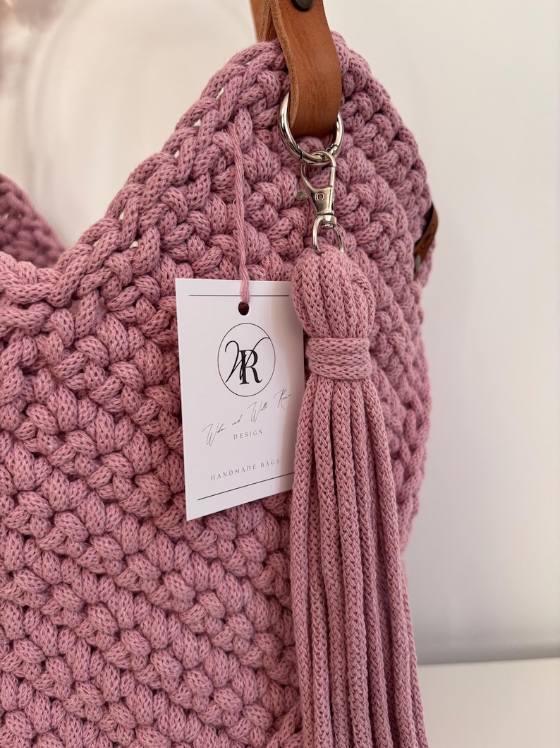 May include: A mauve crocheted handbag featuring a thick, textured knit pattern. The bag includes a brown leather strap attached with a silver metal clasp. A long, matching mauve cord tassel hangs from the clasp, and a white rectangular tag with the logo WR and the text "Willa and Willa Rose Design Handmade Bags" is attached.