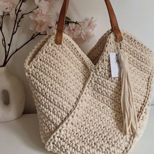 K&ouml;nnte beinhalten: Eine cremefarbene H&auml;keltasche mit einem dicken, strukturierten Strickmuster, zwei braunen Leder-Schulterriemen, die mit Metallnieten befestigt sind, und einer passenden cremefarbenen Quaste, die an einem Metallring mit einem kleinen wei&szlig;en Etikett h&auml;ngt.