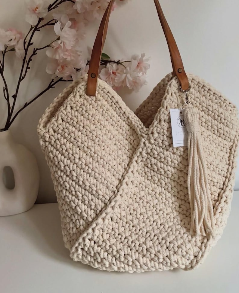 K&ouml;nnte beinhalten: Eine cremefarbene H&auml;keltasche mit einem dicken, strukturierten Strickmuster, zwei braunen Leder-Schulterriemen, die mit Metallnieten befestigt sind, und einer passenden cremefarbenen Quaste, die an einem Metallring mit einem kleinen wei&szlig;en Etikett h&auml;ngt.