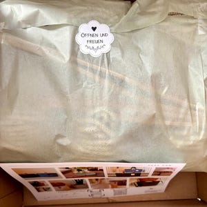 K&ouml;nnte beinhalten: Ein Versandkarton mit in hellgr&uuml;nem Seidenpapier eingewickelten Artikeln, verschlossen mit einem wei&szlig;en, gewellten Aufkleber mit Herzsymbol und dem Text &Ouml;FFNEN UND FREUEN. Unten liegt eine rechteckige Produktkarte mit mehreren kleinen Abbildungen.