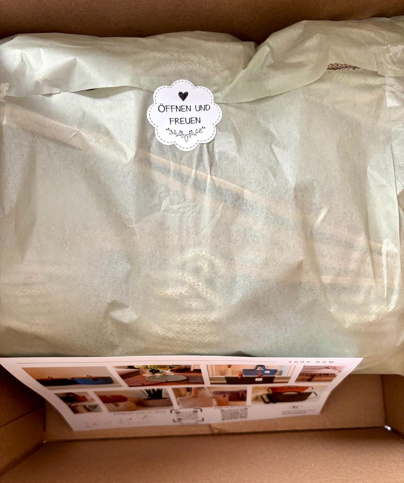 K&ouml;nnte beinhalten: Ein Versandkarton mit in hellgr&uuml;nem Seidenpapier eingewickelten Artikeln, verschlossen mit einem wei&szlig;en, gewellten Aufkleber mit Herzsymbol und dem Text &Ouml;FFNEN UND FREUEN. Unten liegt eine rechteckige Produktkarte mit mehreren kleinen Abbildungen.