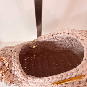 Op de afbeelding: Een beige gehaakte handtas met een dikke gevlochten rand, een goudkleurige metalen magnetische sluiting en een goudkleurige ring aan de zijkant versierd met kleine witte parelachtige kralen die aan een korte gouden ketting hangen.