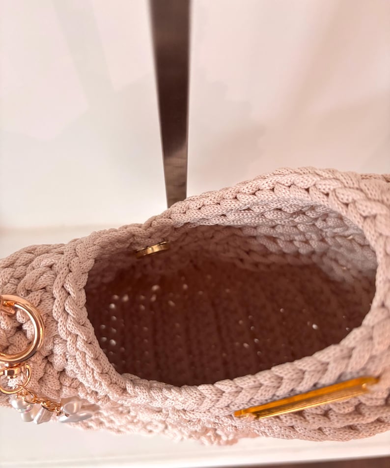 Op de afbeelding: Een beige gehaakte handtas met een dikke gevlochten rand, een goudkleurige metalen magnetische sluiting en een goudkleurige ring aan de zijkant versierd met kleine witte parelachtige kralen die aan een korte gouden ketting hangen.