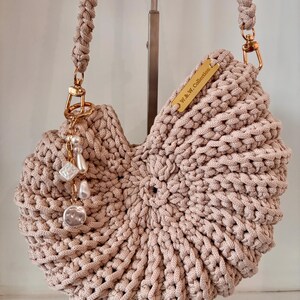 Op de afbeelding: Een beige gehaakte handtas met een cirkelvormig, schelpachtig ontwerp en een gevlochten schouderband. Het heeft goudkleurige hardware, een metalen label met de inscriptie W.&W. Collection en een bedel met witte parelachtige kralen.