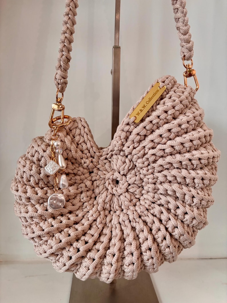 Op de afbeelding: Een beige gehaakte handtas met een cirkelvormig, schelpachtig ontwerp en een gevlochten schouderband. Het heeft goudkleurige hardware, een metalen label met de inscriptie W.&W. Collection en een bedel met witte parelachtige kralen.