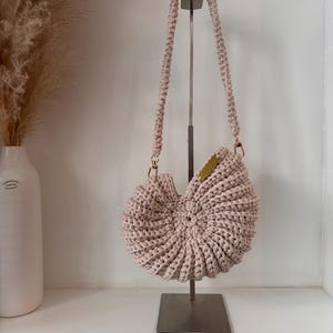 Op de afbeelding: Een lichtbeige gehaakte handtas met een opvallende spiraalvormige schelpvorm. De tas heeft een dikke, gevlochten schouderband bevestigd met goudkleurige metalen hardware en een klein rechthoekig goudkleurig metalen label aan de rechterbovenrand.